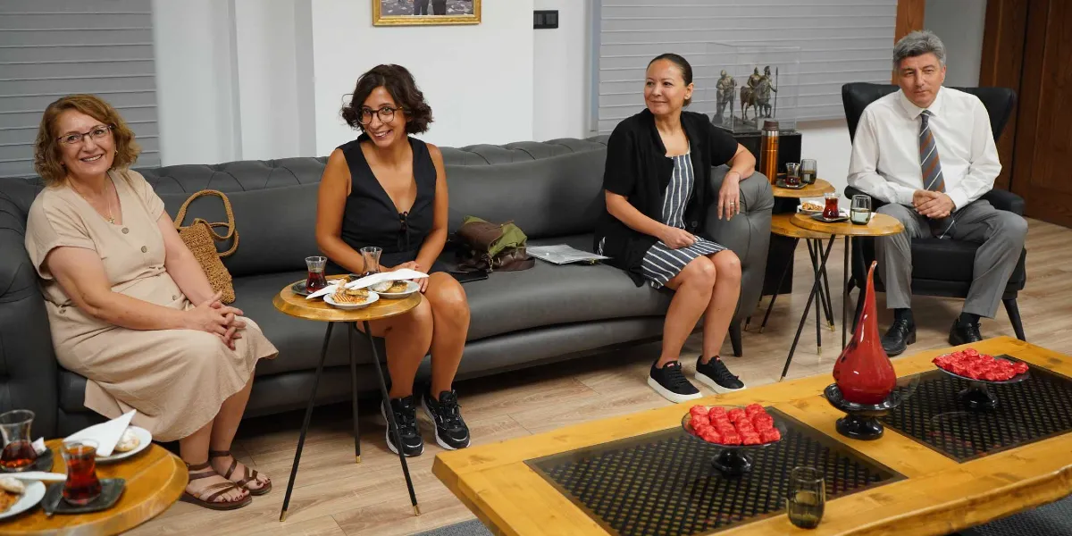 Modern bir ofis ortamında, gri bir koltukta yan yana oturan üç kadın ve yanlarındaki tekli koltukta oturan bir erkek, sohbet ederken gülümseyerek kameraya bakıyor. Önlerinde çay ve atıştırmalıkların bulunduğu ahşap sehpalar yer alıyor. Kadınlar günlük kıyafetler, erkek ise beyaz gömlek ve kravat giyiyor.