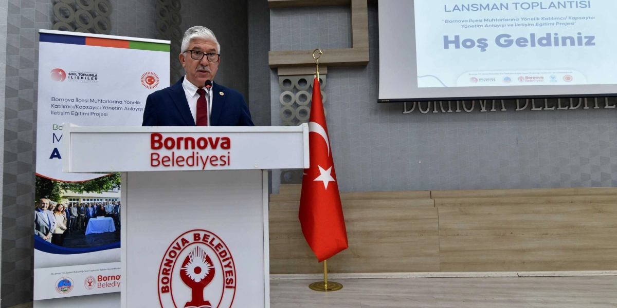 Gri saçlı, gözlüklü, lacivert ceket ve kırmızı kravatlı bir yetkili, üzerinde Bornova Belediyesi logosu bulunan kürsüde konuşma yapıyor. Kürsünün yanında Türk bayrağı, arkasında ise üzerinde 