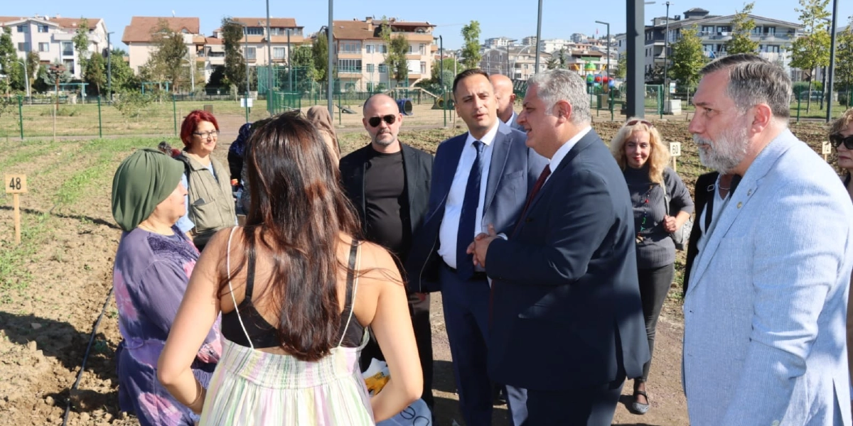 Açık havada, toprak bir alanda bir araya gelmiş bir grup insan, Bornova'daki bir tarım projesini inceliyor. Ön planda sırtı dönük, askılı bir elbise giyen bir kadın ve yanında yeşil başörtülü bir kadın bulunuyor. Onların karşısında, takım elbiseli ve resmi giyimli erkeklerin de aralarında olduğu bir grup, sohbet ederek alanı gözlemliyor. Arka planda ise üzerinde numaralar yazılı küçük tabelaların bulunduğu ekili alanlar, çitler ve yerleşim yerleri görülüyor.