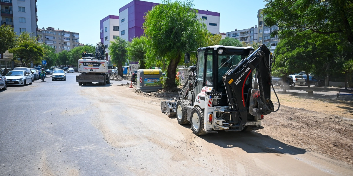 Bornova merkezinde bir cadde üzerinde devam eden yol yenileme çalışmaları kapsamında, biri ön planda diğeri ileride olmak üzere iki adet Hidromek marka iş makinesi çalışıyor. Asfaltın bir kısmının kazındığı caddede, ağaçların ve park halindeki araçların bulunduğu çevrede belediye ekiplerinin yürüttüğü düzenleme çalışmaları görülüyor.