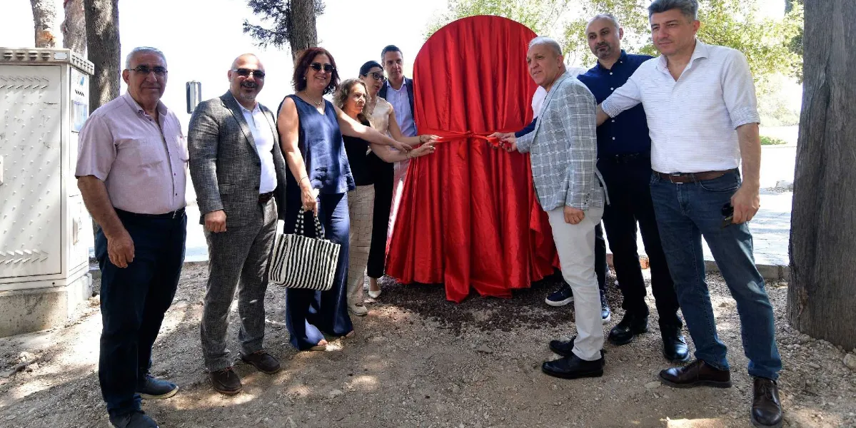 Açık havada, ağaçlık bir alanda bir araya gelmiş bir grup insan, kırmızı bir örtüyle kaplı anıtın açılışını yapmak için kurdeleyi kesiyor. Gruptaki kişiler şık kıyafetler içinde yan yana dururken, bir kısmı ellerini kırmızı kurdelenin üzerinde tutuyor. Arka planda, üzerinde kırmızı örtü bulunan dairesel formdaki anıt dikkat çekiyor.