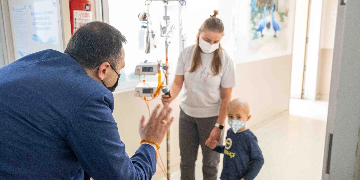 Mavi ceketli ve maskeli bir erkek, hastane koridorunda maske takan küçük bir çocuğa ve ona eşlik eden maskeli bir kadına el sallayarak selam veriyor. Küçük çocuk, yanında serum askısı bulunan annesinin elini tutuyor.