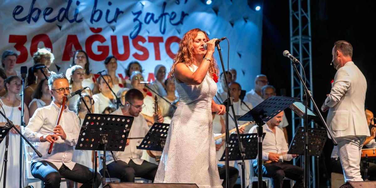30 Ağustos Zafer Bayramı kutlamaları kapsamında düzenlenen konserde, beyaz kıyafetli bir kadın solist mikrofon başında şarkı söylüyor. Sahne arkasındaki dev pankartta 