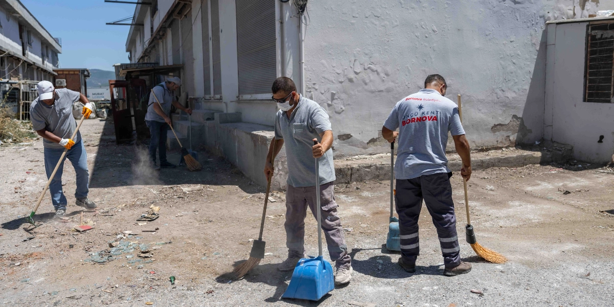 Bornova Belediyesi logolu gri tişörtler giyen dört temizlik işçisi, sanayi tipi bir binanın önündeki boş alanda süpürgelerle temizlik çalışması yapıyor. İşçilerden biri maske takarken, diğerleri ellerindeki süpürge ve faraşlarla yerdeki atıkları topluyor.