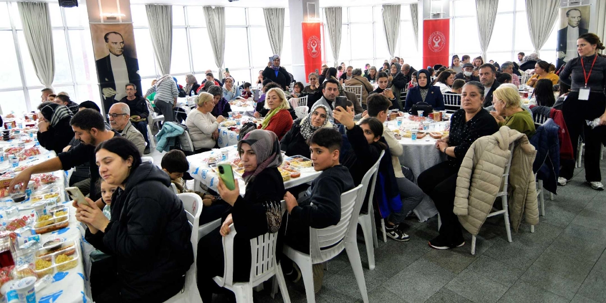 Geniş ve aydınlık bir salonda düzenlenen toplu yemek etkinliğinde, beyaz örtülü masaların etrafında oturan çok sayıda Bornovalı vatandaş yer alıyor. Katılımcılar önlerindeki yemek paketleriyle ilgilenirken, bazıları cep telefonlarıyla fotoğraf çekiyor. Salonun duvarlarında Atatürk portreleri ve Bornova Belediyesi'nin logolu kırmızı flamaları asılı bulunuyor. Sağ tarafta ayakta duran görevli bir kadın, elindeki yaka kartıyla etkinliği takip ediyor.