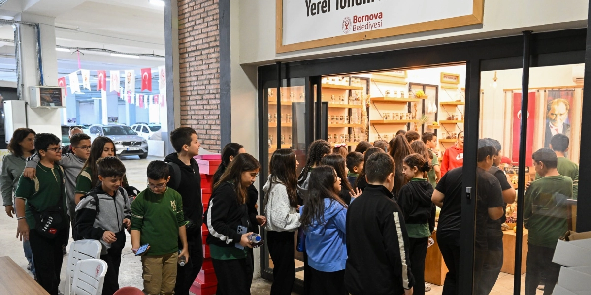 Bornova Belediyesi'ne ait Yerel Tohum Merkezi'nin önünde toplanmış, içerideki tohum koleksiyonunu inceleyen bir grup öğrenci. Çocuklar, cam kapılı merkezin önünde sıraya girmiş veya içeriyi gözlemliyor; merkezin içindeki raflarda cam kavanozlar içinde tohumlar sergileniyor ve duvarda Atatürk posteri asılı.
