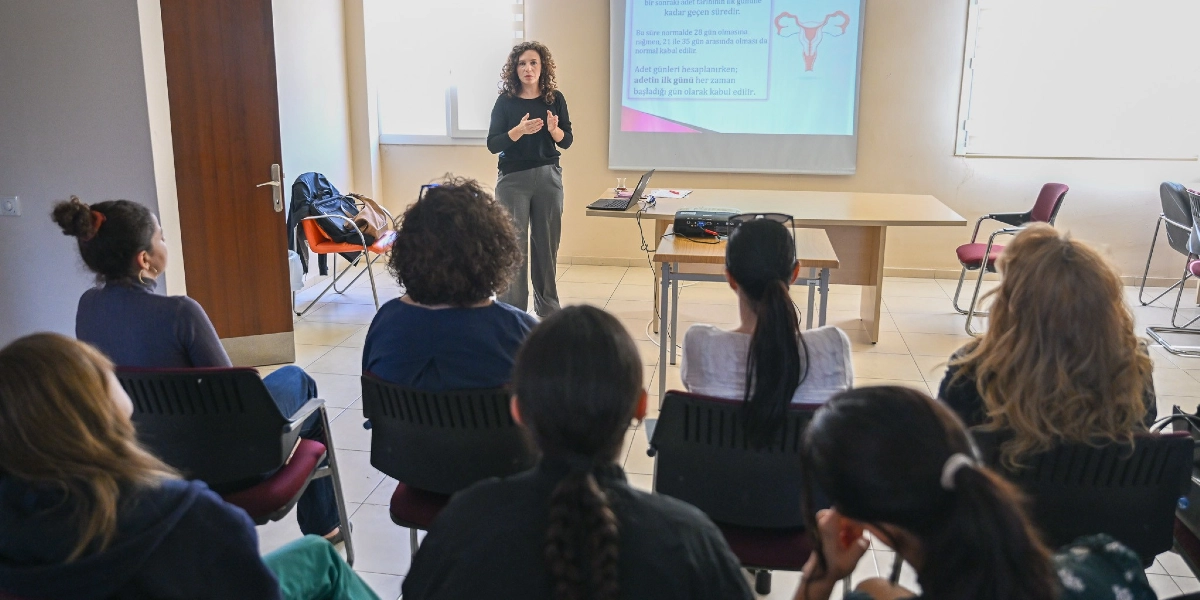 Kıvırcık saçlı bir kadın, bir sınıfta kadın sağlığı konulu sunum yaparken katılımcılara hitap ediyor. Arkasındaki projeksiyon perdesinde adet döngüsü ve hesaplanması hakkında bilgiler ile rahim anatomisini gösteren bir şema yer alıyor. Katılımcılar ise sıralarda oturmuş, konuşmacıyı dikkatle dinliyor.