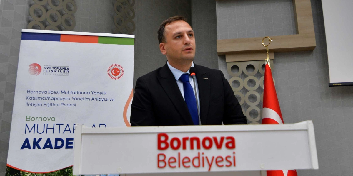 Koyu renk takım elbiseli ve mavi kravatlı bir erkek, üzerinde Bornova Belediyesi yazan bir kürsüde mikrofon başında konuşma yapıyor. Arka planda, üzerinde T.C. İçişleri Bakanlığı Sivil Toplumla İlişkiler Genel Müdürlüğü logosu ile 
