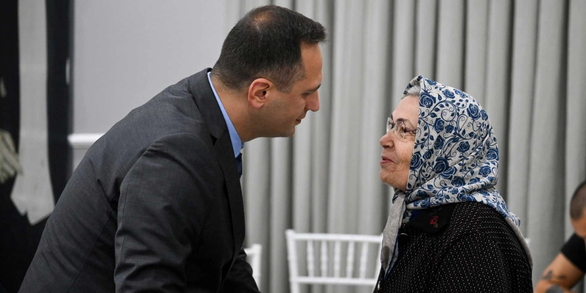 Koyu renk takım elbiseli bir erkek, başörtülü yaşlı bir kadınla samimi bir şekilde sohbet ediyor. İkili birbirine bakarak gülümsüyor.