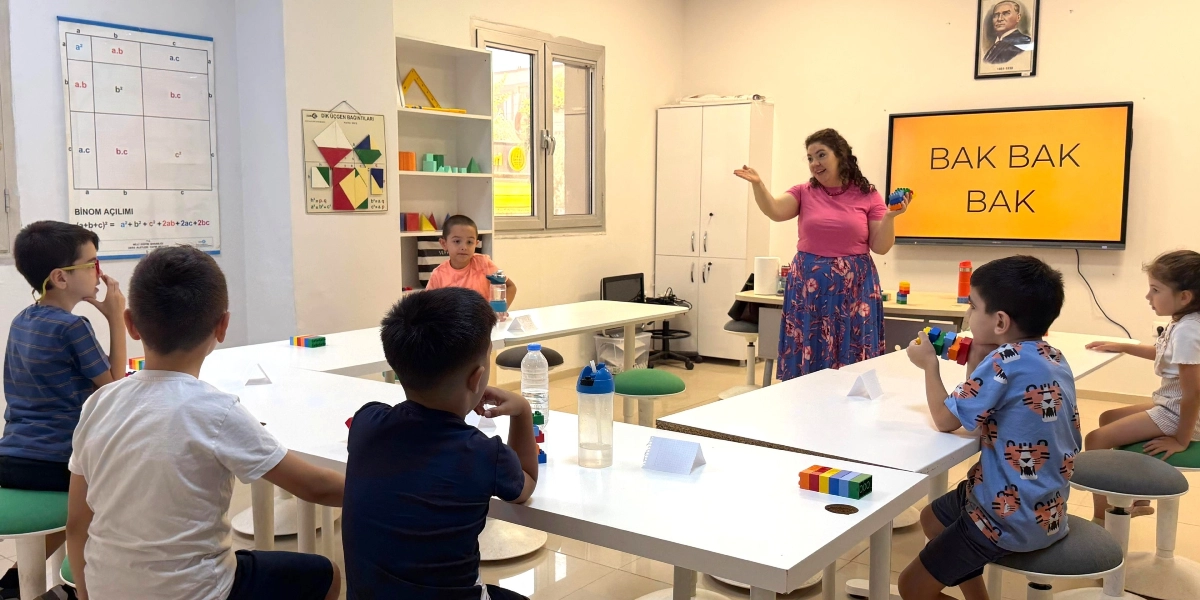 Sınıf ortamında, pembe tişörtlü bir öğretmen elinde tuttuğu renkli plastik tuğlalarla öğrencilerine eğitim veriyor. Öğrenciler, U şeklindeki masalarda oturmuş, önlerinde bulunan benzer renkli tuğlalarla çalışıyorlar. Arkadaki akıllı tahtada büyük harflerle 