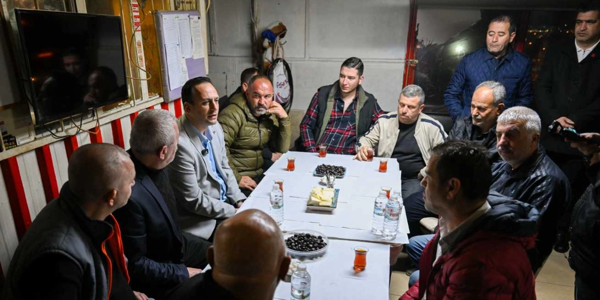 Kapalı bir mekanda, beyaz örtülü bir masa etrafında toplanmış on bir kişilik erkek grubu, sabahın erken saatlerinde kahvaltı yapıyor. Masanın ortasında zeytin ve peynir tabakları, su şişeleri ve ince belli bardaklarda çaylar bulunuyor. Gruptakiler günlük kıyafetler içinde, bir kısmı otururken bir kısmı ayakta duruyor ve birbirleriyle sohbet ediyor.
