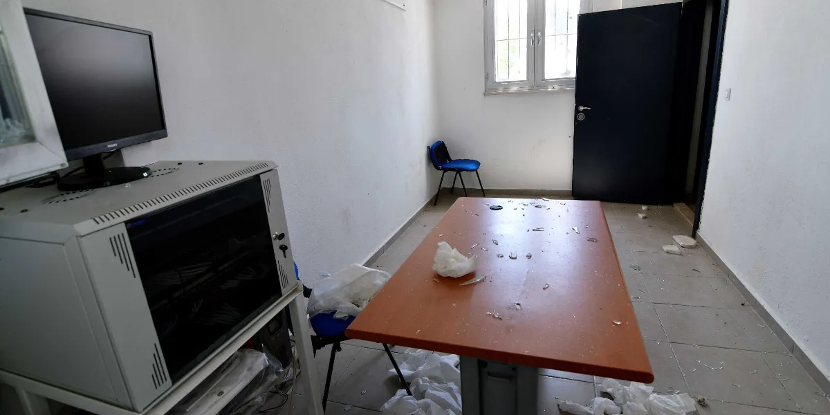 Beyaz duvarlı, sade bir ofis odasında, ahşap bir çalışma masasının üzeri ve yerler kırık cam parçalarıyla dolu. Masanın üzerinde buruşturulmuş kağıtlar ve cam kırıkları bulunurken, odanın köşesinde tek bir mavi sandalye, masanın yanında ise başka bir mavi sandalye ve üzerinde bir bilgisayar monitörü bulunan bir sunucu kabini yer alıyor. Odanın arka kısmında siyah bir kapı ve dışarıya açılan pencereli bir duvar bulunuyor.