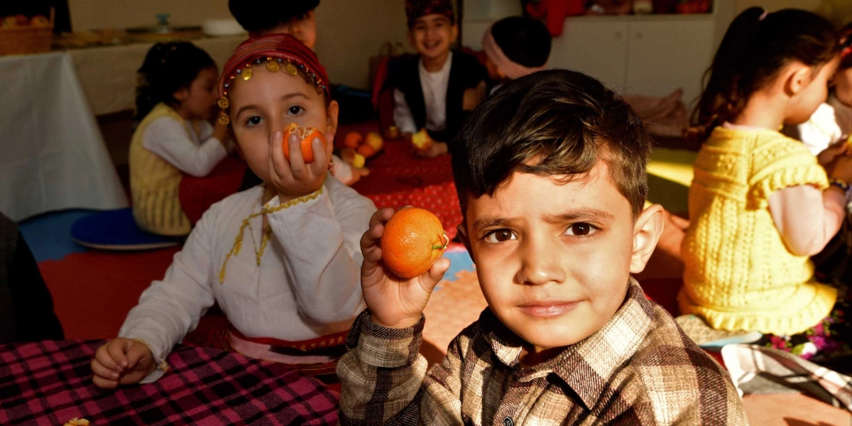Yerli Malı Haftası etkinliğinde, geleneksel kıyafetler ve günlük kıyafetler içindeki çocuklar masalarda oturuyor. Ön planda ekose gömlekli bir erkek çocuk elinde bir mandalina tutarak kameraya bakıyor; hemen yanında ise başında altın işlemeli bir başlık bulunan bir kız çocuğu elindeki mandalinayı yiyor. Arka planda ise diğer çocuklar etkinliklerine devam ediyor.