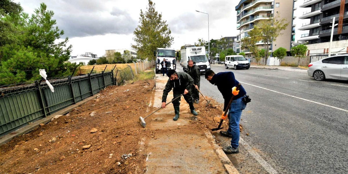 Belediye işçileri, Bornova'da bir yol kenarındaki toprak zemini ve kaldırımı temizleyerek düzenleme çalışması yapıyor. İşçilerden biri süpürgeyle toprakları süpürürken, diğeri kürekle çalışıyor; arka planda ise trafik akışının olduğu asfalt bir yol ve modern apartmanlar görülüyor.