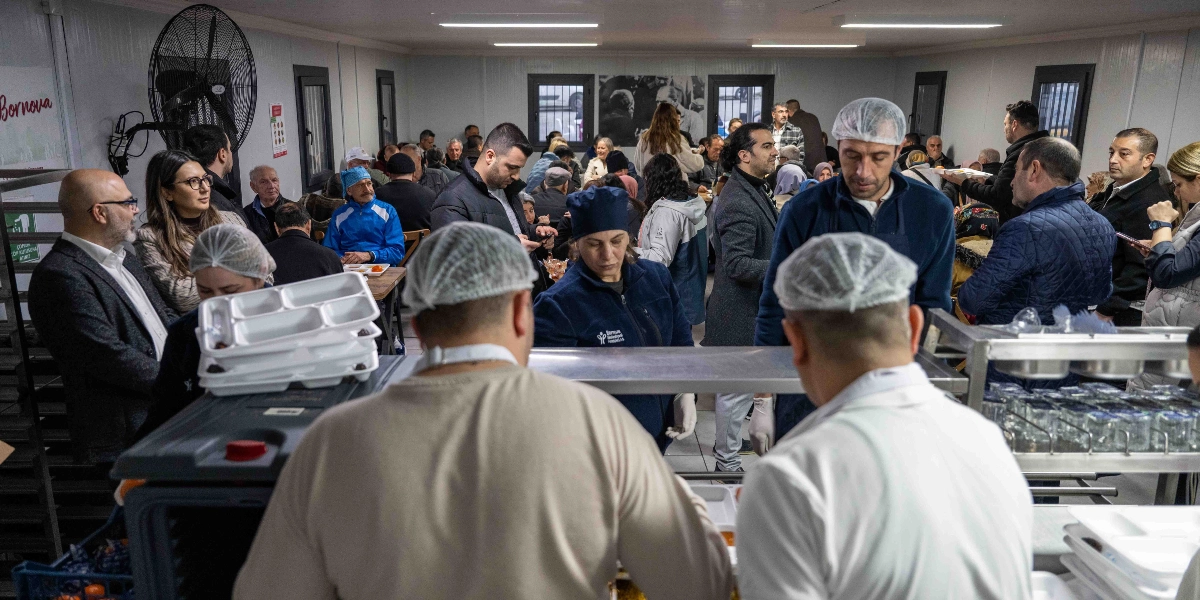 Kapalı bir yemekhanede, beyaz önlük ve bone giymiş görevlilerin servis yaptığı, kalabalık bir grubun iftar yemeği için bir araya geldiği ortam. Ön planda servis yapan iki görevli, arka planda ise masalarda oturan ve yemek bekleyen çok sayıda insan görülüyor.