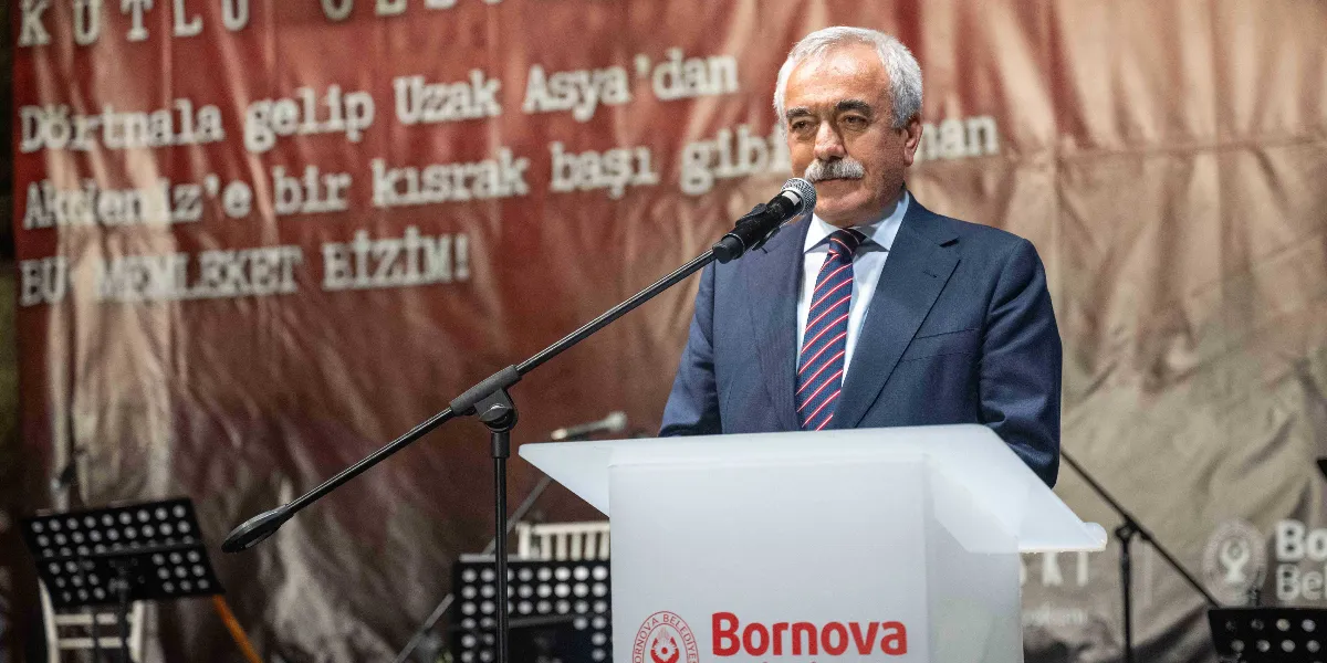 Bornova Belediye Başkanı Ömer Eşi, üzerinde Bornova Belediyesi logosu bulunan bir kürsüde, mikrofon başında konuşma yapıyor. Arka planda, üzerinde Nazım Hikmet’in 