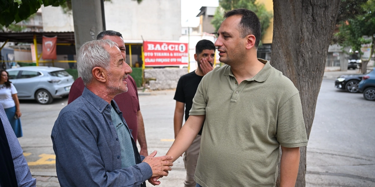 Bornova'da bir sokakta, haki renkli polo yaka tişört giyen bir erkek, yaşlı bir adamla tokalaşarak sohbet ediyor. Arka planda başka erkekler ve park halindeki araçlar bulunurken, bir işletmenin üzerinde 