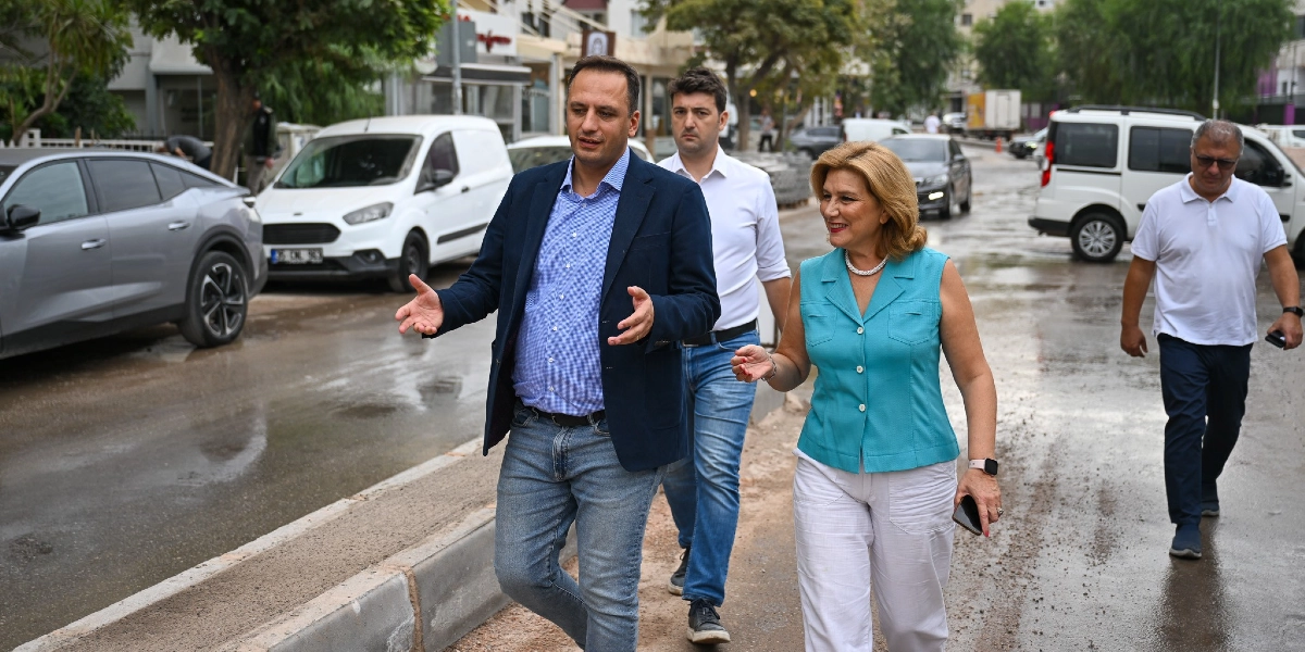 Bornova Zafer Caddesi'nde incelemelerde bulunan bir grup, cadde üzerinde yürürken görülüyor. Önde, lacivert ceketli ve mavi gömlekli bir erkek ile yanında turkuaz kolsuz bluz ve beyaz pantolon giyen bir kadın, konuşarak ilerliyor. Arkalarında beyaz gömlekli bir erkek ve biraz daha geride beyaz tişörtlü başka bir erkek daha bulunuyor. Arka planda park halindeki araçlar ve cadde düzenleme çalışmaları göze çarpıyor.