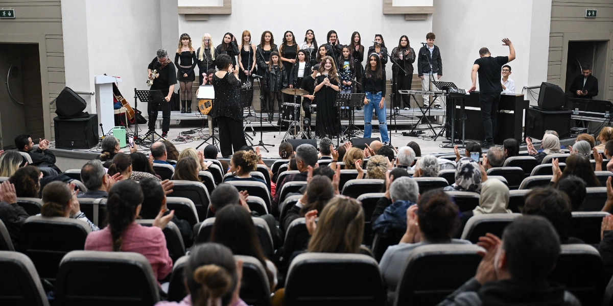 Modern bir konferans salonunda, sahnede müzik aletleri eşliğinde şarkı söyleyen gençlerden oluşan bir koro ve onları yöneten bir şef yer alıyor. Siyah kıyafetler giymiş genç kızlar ve erkeklerden oluşan koro üyeleri, mikrofonlar önünde performans sergiliyor. Sahnenin solunda gitar çalan bir müzisyen, sağında ise piyano başında oturan bir kişi bulunuyor. Salonu dolduran izleyiciler, sahnedeki performansı alkışlarla izliyor.