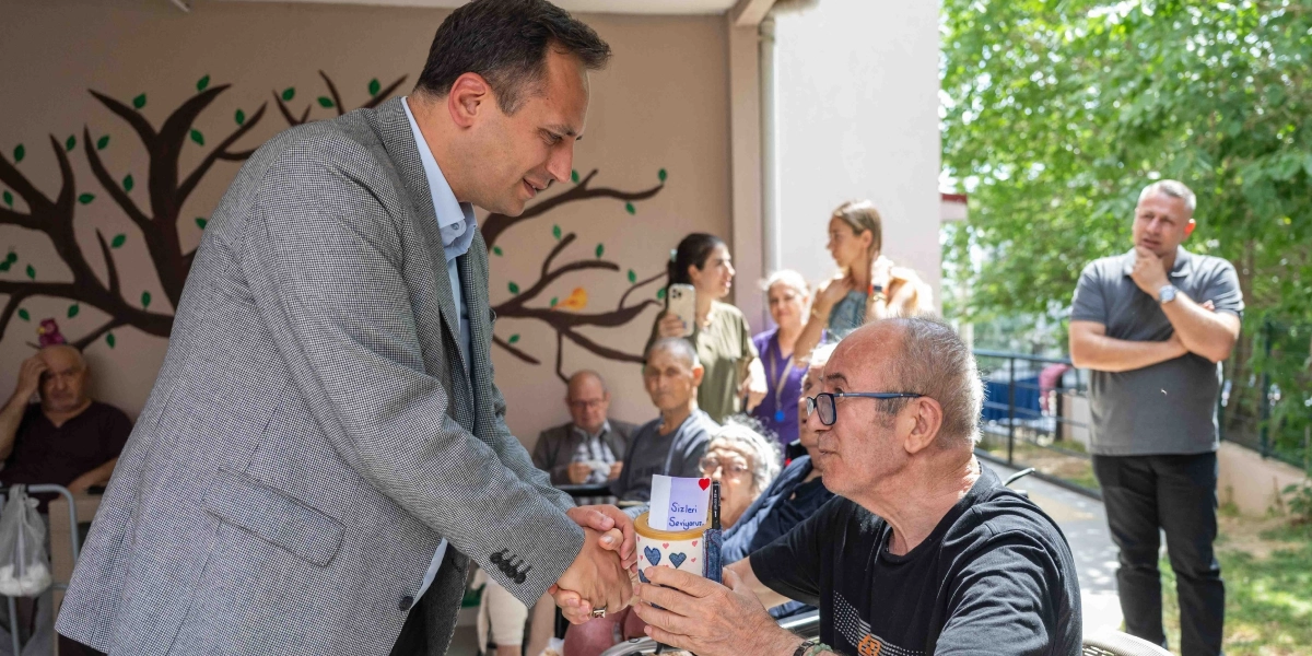 Gri ceketli bir erkek, Babalar Günü etkinliğinde yaşlı bir adamın elini sıkıyor. Yaşlı adam, üzerinde 