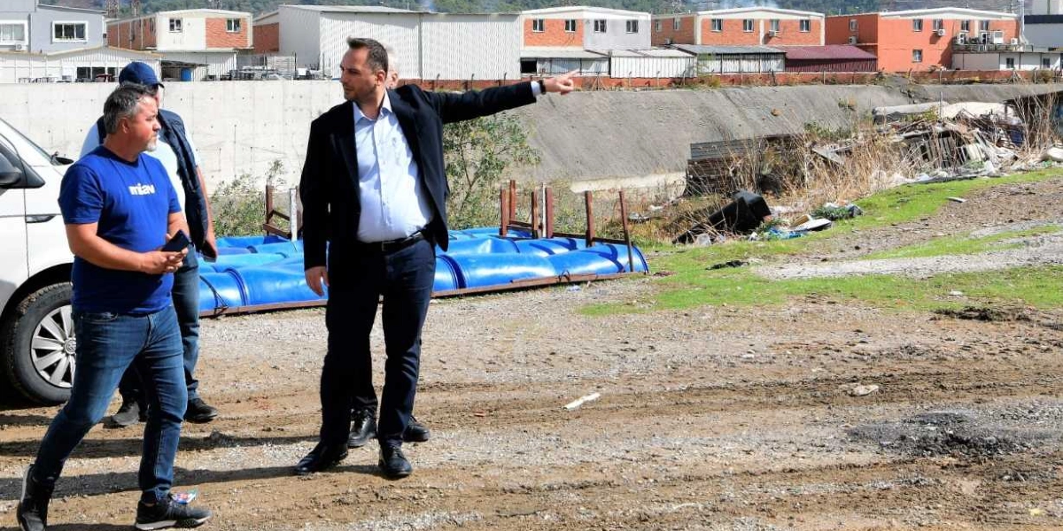 Açık havada, toprak bir alanda duran iki erkekten biri, üzerinde mavi tişört ve kot pantolonla yanında dururken, diğeri lacivert ceket ve pantolon giymiş bir şekilde eliyle ilerideki boş araziyi işaret ediyor. Arka planda mavi plastik variller ve sanayi tipi binalar yer alıyor.