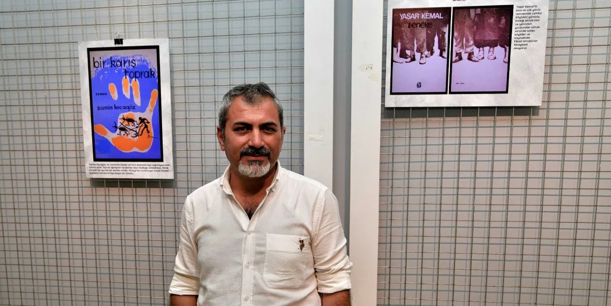 Beyaz gömlekli, kirli sakallı bir adam, üzerinde edebiyat eserlerinin tanıtım afişlerinin asılı olduğu metal bir ızgara panonun önünde duruyor. Sol taraftaki afişte Samim Kocagöz'ün 