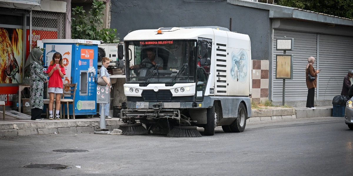 Bornova Belediyesi'ne ait beyaz bir yol süpürme aracı, cadde üzerinde temizlik çalışması yapıyor. Aracın ön kısmında 