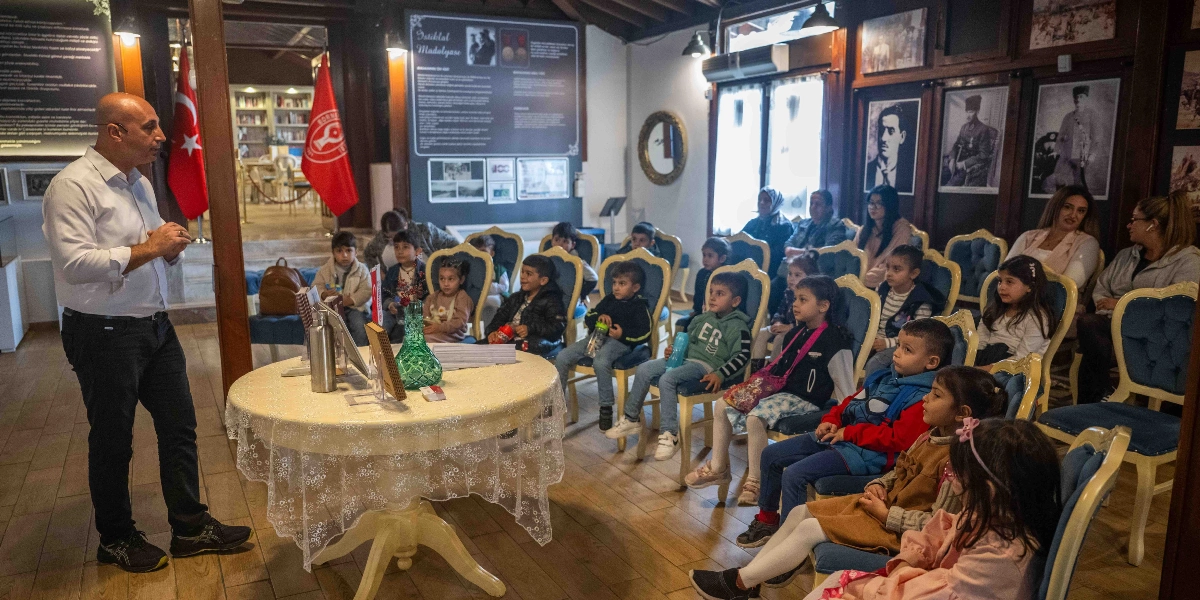 Belkahve'deki tarihi bir mekanda, beyaz gömlekli bir erkek ayakta durarak karşısında oturan çocuk ve kadınlardan oluşan gruba konuşma yapıyor. Ortada dantel örtülü yuvarlak bir masa, arka planda ise üzerinde 