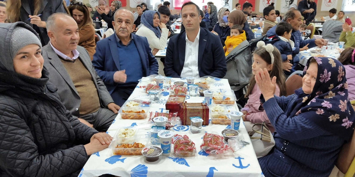 Bornova Belediye Başkanı Ömer Eşki, bir yemek masasında vatandaşlarla birlikte oturuyor. Masanın üzerinde paketli yemekler, ayranlar ve ekmekler bulunuyor. Başkan Eşki'nin sağında ve solunda çeşitli yaş gruplarından vatandaşlar yer alırken, masanın sağ ucunda oturan başörtülü yaşlı bir kadın ellerini çırparak gülümsüyor. Arka planda ise kalabalık bir yemek salonu ve masalar görülüyor.