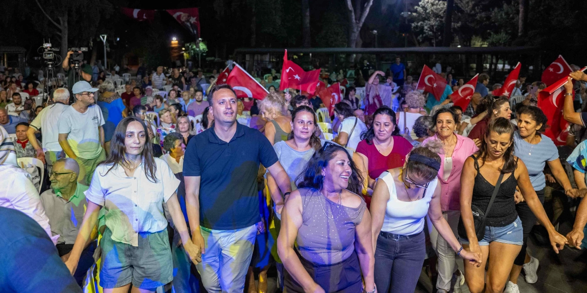 Gece saatlerinde açık havada düzenlenen bir etkinlikte, ellerinde Türk bayrakları tutan kalabalık bir grup, el ele tutuşarak halay çekiyor. Ön planda, beyaz gömlekli bir kadın, lacivert polo yaka tişörtlü bir erkek ve yanında neşeyle gülümseyen diğer katılımcılar yer alıyor. Arka planda ise etkinliği izleyen çok sayıda kişi ve sahneyi kaydeden bir kamera ekibi bulunuyor.