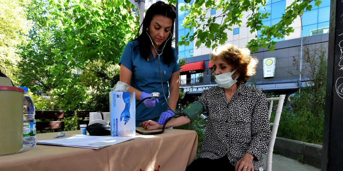 Açık havada, ağaçların altında kurulan bir sağlık standında, mavi üniformalı bir sağlık çalışanı yaşlı bir kadının tansiyonunu ölçüyor. Sağlık çalışanı stetoskop kullanarak kadının koluna takılı tansiyon aletini kontrol ederken, maske takan yaşlı kadın oturur vaziyette bekliyor. Masanın üzerinde bir kutu eldiven ve su şişesi bulunuyor.