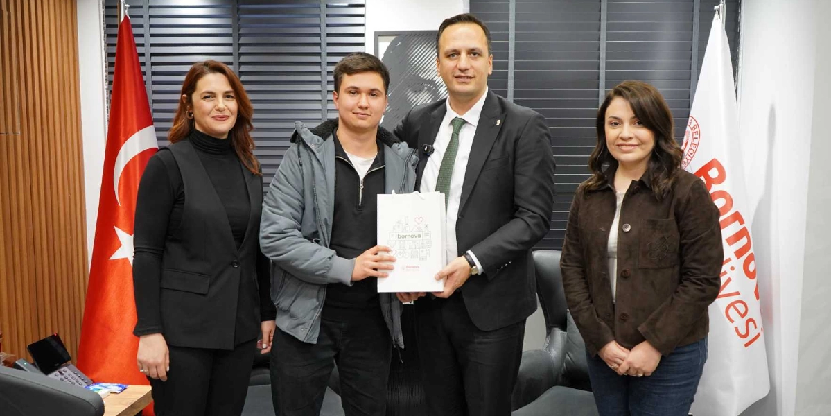 Bornova Belediye Başkanı, yanında duran genç bir öğrenciye üzerinde Bornova Belediyesi logosu bulunan beyaz bir hediye çantası takdim ediyor. Yanlarında iki kadın yetkili daha bulunuyor. Arka planda Türk bayrağı ve Bornova Belediyesi flaması yer alıyor.