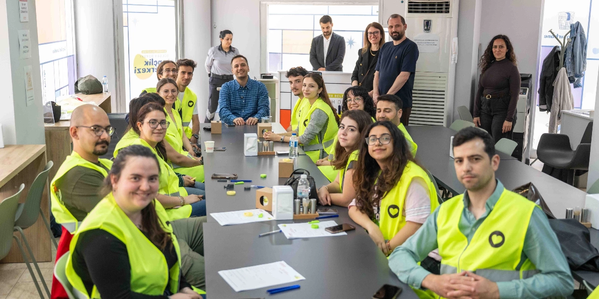 Bornova'da bir ofis ortamında, üzerinde kalp figürü bulunan sarı yelekler giymiş gençlerden oluşan bir grup, uzun bir toplantı masasının etrafında oturuyor. Katılımcıların çoğu masanın iki yanında karşılıklı yer alırken, bazıları ayakta duruyor. Masanın üzerinde kağıtlar, kalemler ve içecekler bulunuyor. Genel atmosfer oldukça samimi ve iş birliği odaklı görünüyor.