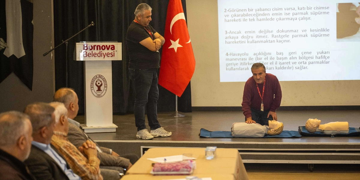 Bornova Belediyesi kürsüsünün yanında duran bir eğitmen, katılımcılara ilk yardım eğitimi veriyor. Eğitmen, sahnede yere serili matlar üzerinde bulunan iki adet ilk yardım mankeni üzerinde kalp masajı ve havayolu açma tekniklerini uygulamalı olarak gösteriyor. Arkadaki projeksiyon perdesinde ise ilk yardım müdahaleleriyle ilgili yazılı talimatlar yer alıyor. Salonun ön sıralarında oturan muhtarlar, eğitimi dikkatle takip ediyor.