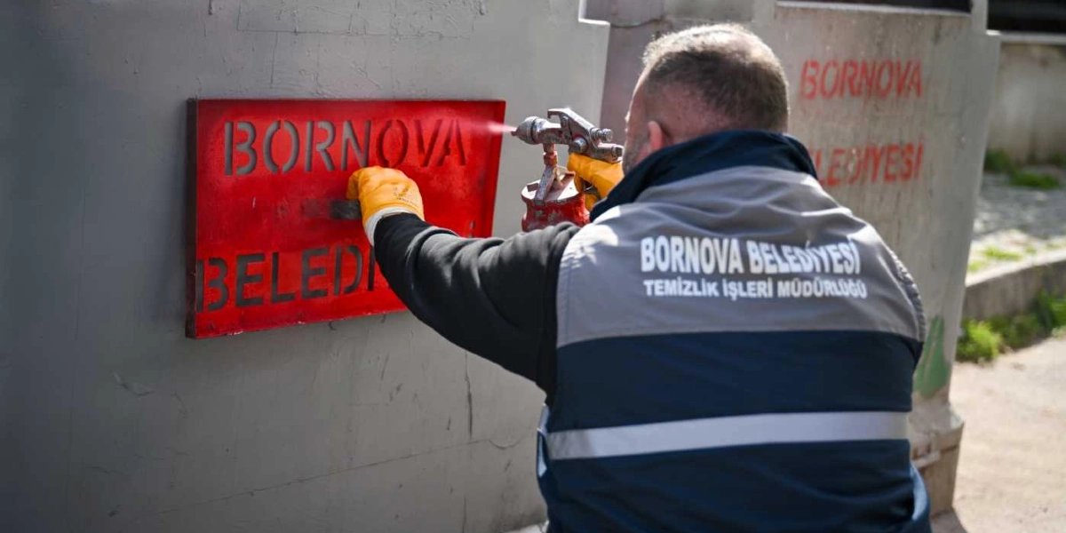 Sırtında Bornova Belediyesi Temizlik İşleri Müdürlüğü yazılı yelek bulunan bir belediye çalışanı, elindeki şablonu bir çöp konteynerine dayayarak sprey boya ile 
