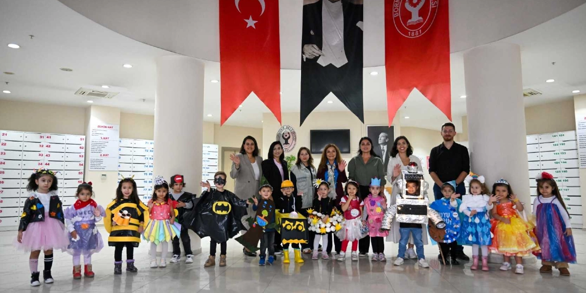 Atık malzemelerden yapılmış geri dönüşüm temalı kostümler giyen bir grup çocuk, arkalarında öğretmenleri ve Bornova Belediyesi yetkilileriyle birlikte kapalı bir alanda poz veriyor. Çocukların kostümleri arasında arı, Batman, robot, çiçek ve çeşitli renkli kıyafetler yer alıyor. Arka planda Türk bayrağı, Atatürk posteri ve Bornova Belediyesi flaması asılı dururken, yanlarda posta kutularını andıran bölmeler bulunuyor.