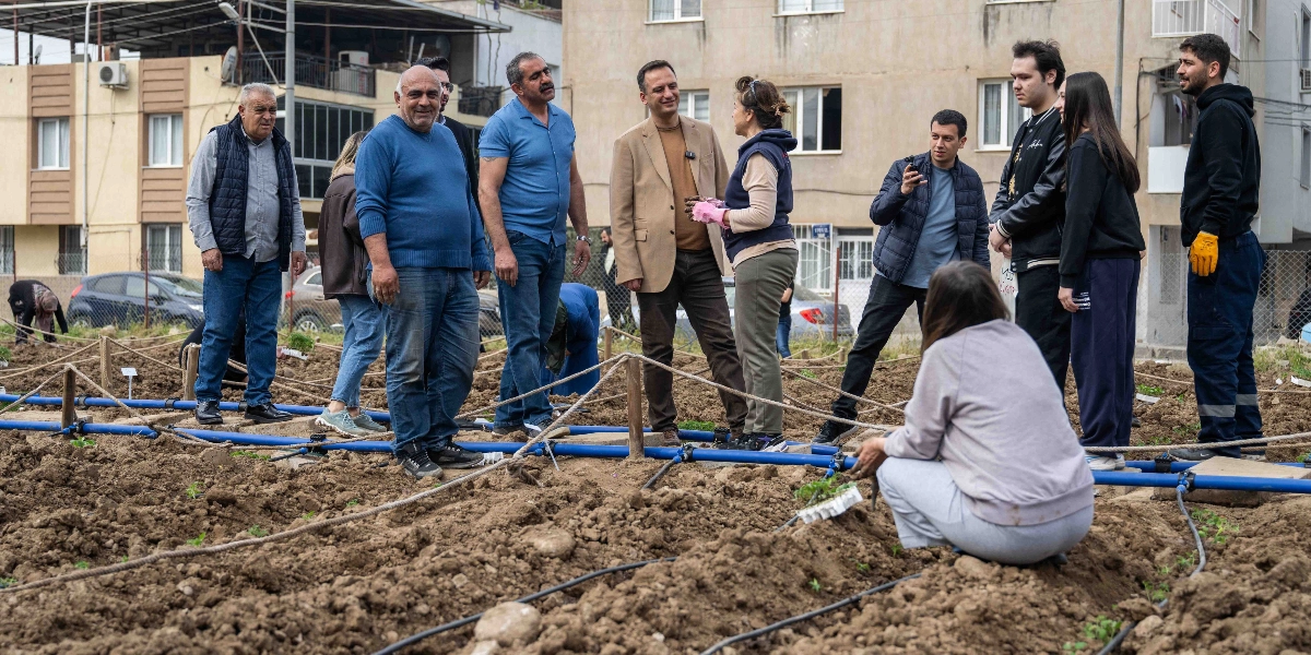 Bornova'da bir kentsel tarım alanında, toprakla uğraşan bir grup insan yer alıyor. Ön planda, toprak üzerinde çömelmiş bir kişi fide dikimi yaparken, arka planda farklı yaş gruplarından insanlar bir arada duruyor. Bir kısmı ayakta sohbet ederken, bazıları da tarım alanındaki sulama boruları ve iplerle çevrili parselleri inceliyor. Arka planda çok katlı apartmanların görüldüğü açık hava ortamında, katılımcılar gündelik kıyafetlerle tarım faaliyetine katılıyor.