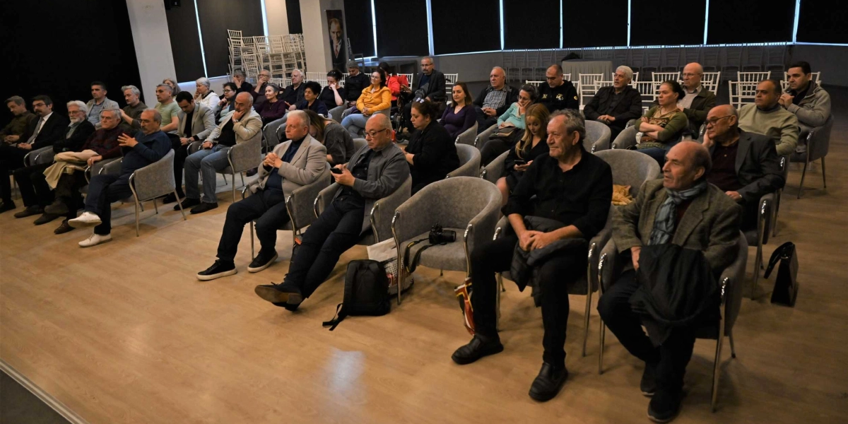 Modern bir konferans salonunda, gri koltuklarda oturan ve dikkatle dinleyen yaklaşık 30 kişilik bir izleyici grubu. Katılımcılar çoğunlukla orta yaş ve üzeri bireylerden oluşuyor; bazıları günlük, bazıları ise daha resmi kıyafetler tercih etmiş. Salonun zemininde açık renkli ahşap parke bulunuyor, arka planda ise üst üste dizilmiş boş sandalyeler ve duvarda Atatürk portresi göze çarpıyor. İzleyiciler sahneye doğru dönük şekilde, yarı dairesel bir düzenle oturuyor.