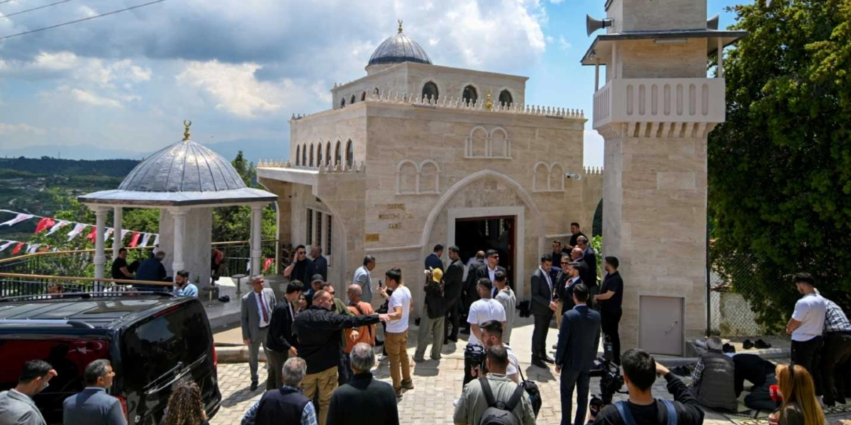 Açık renkli taşlardan inşa edilmiş, kubbeli ve minareli yeni bir caminin önünde toplanmış kalabalık bir grup insan görülüyor. Caminin girişinde ve çevresinde takım elbiseli protokol üyeleri, basın mensupları ve vatandaşlar yer alıyor. Caminin sol tarafında, çatısı kubbeli, sütunlu küçük bir yapı ve çevresinde asılı Türk bayrakları bulunuyor. Arka planda ise ağaçlık bir arazi ve açık bir gökyüzü seçiliyor.