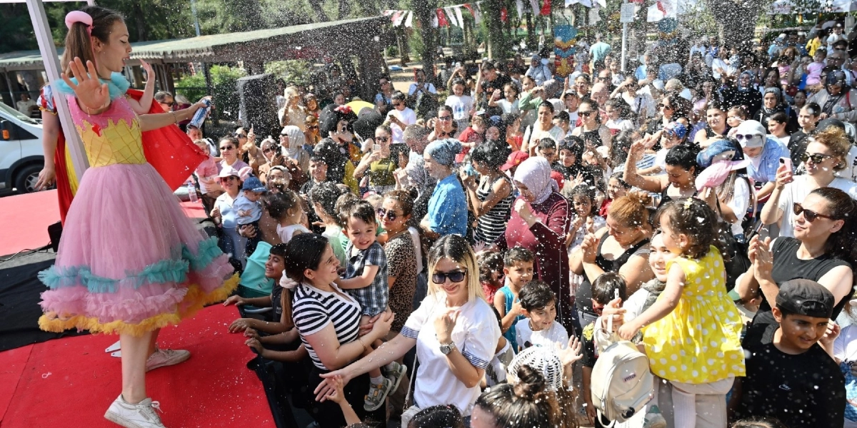 Bornova Büyükpark'ta düzenlenen bir etkinlikte, sahne üzerinde renkli kostümlü bir animatörün çocukların üzerine köpük sıktığı, çocukların ve ailelerinin eğlenceli anlar yaşadığı kalabalık bir açık hava etkinliği.