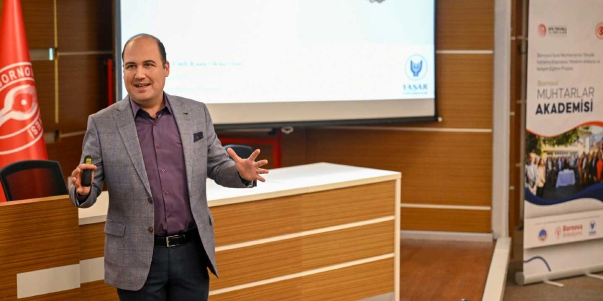 Gri ekose ceket ve mor gömlek giymiş bir konuşmacı, bir toplantı salonunda ellerini kullanarak sunum yapıyor. Arkasında projeksiyon perdesi ve sağ tarafta üzerinde 