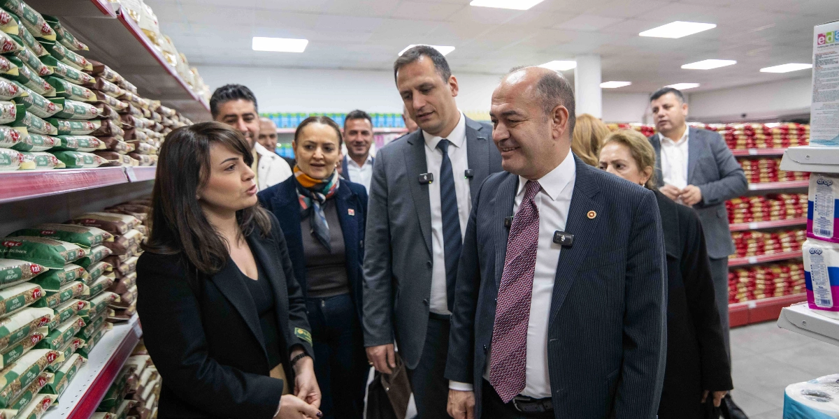 CHP Genel Başkan Yardımcısı Özgür Karabat ve beraberindeki heyet, bir marketin gıda reyonları arasında incelemelerde bulunuyor. Karabat, takım elbiseli ve kravatlı bir şekilde gruptakilerle sohbet ederken, yanında bulunan diğer yetkililer de market raflarındaki ürünleri inceliyor. Arka planda market çalışanları ve diğer heyet üyeleri yer alıyor.