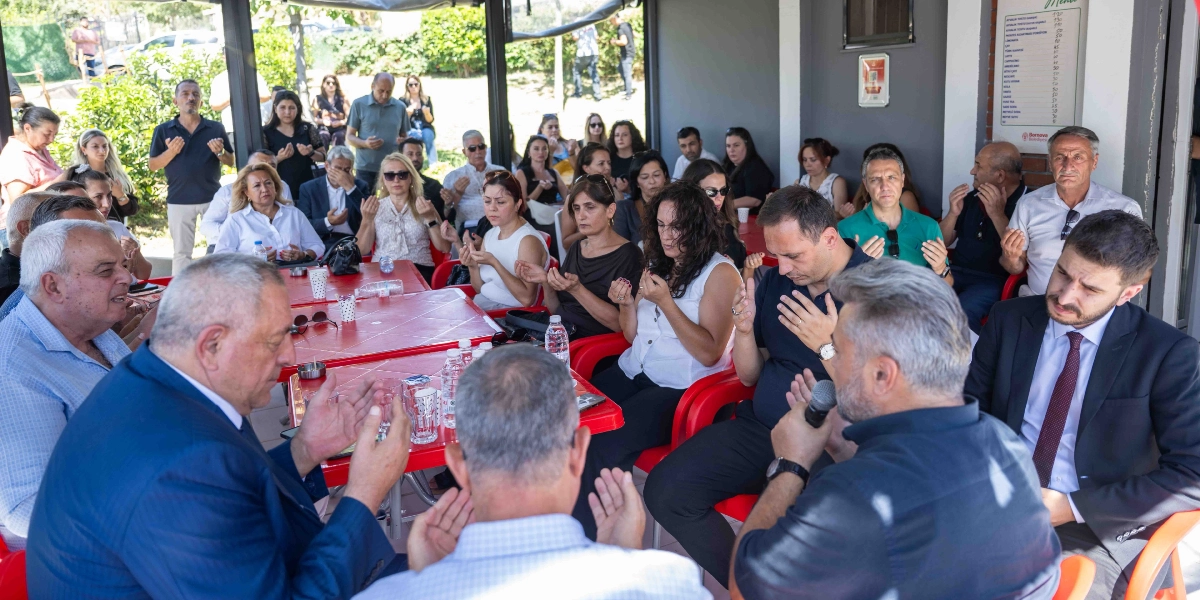 Açık havada, masalar etrafında toplanmış kalabalık bir grup insan, ellerini açarak dua ediyor. Önde, lacivert ceketli bir adam ve yanında mikrofon tutan başka bir kişi dikkat çekerken, arkada oturan kadınlar ve erkekler de aynı şekilde dua ediyor. Ortamda masaların üzerinde su şişeleri ve bardaklar bulunuyor, arka planda ise Bornova Belediyesi logosunun yer aldığı bir menü panosu görünüyor.