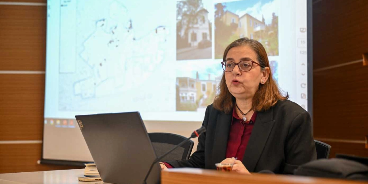 Gözlüklü, kahverengi saçlı bir kadın, önünde dizüstü bilgisayar ve mikrofon bulunan bir masada oturarak konuşma yapıyor. Arka plandaki projeksiyon perdesinde Bornova'daki tarihi yapıları gösteren fotoğraflar ve bir harita taslağı yer alıyor.