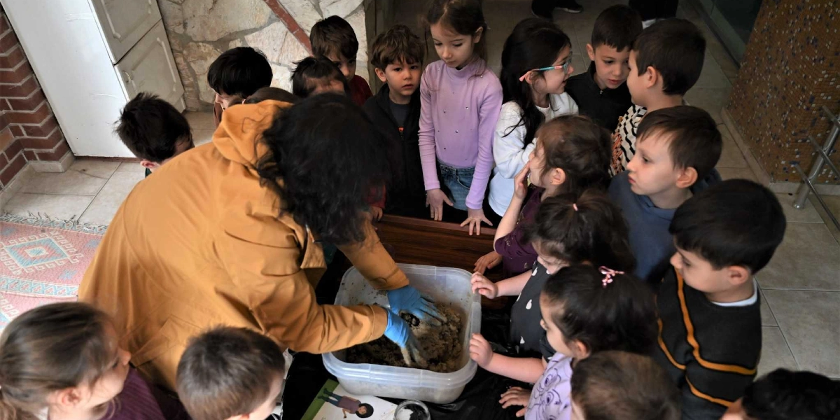 Sarı ceketli bir eğitmen, plastik eldivenler takarak şeffaf bir kap içerisindeki toprak karışımını hazırlarken, etrafına toplanmış bir grup anaokulu öğrencisi merakla onu izliyor.