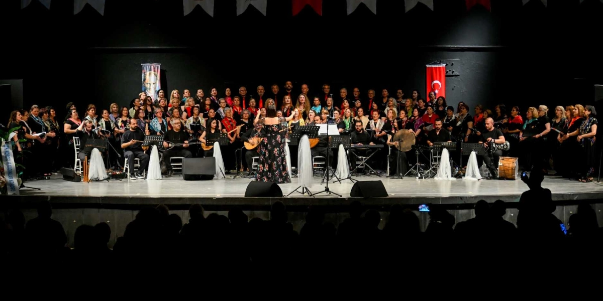 Sahne üzerinde siyah kıyafetler giymiş geniş bir koro ve müzisyen grubu, ellerinde kaşıklar ve çeşitli enstrümanlarla performans sergiliyor. Koronun önünde orkestra şefi sırtı dönük şekilde dururken, arka planda Atatürk posteri ve Türk bayrağı asılı. Sahne ışıklandırılmış, ön planda ise karanlıkta izleyicilerin silüetleri görülüyor.
