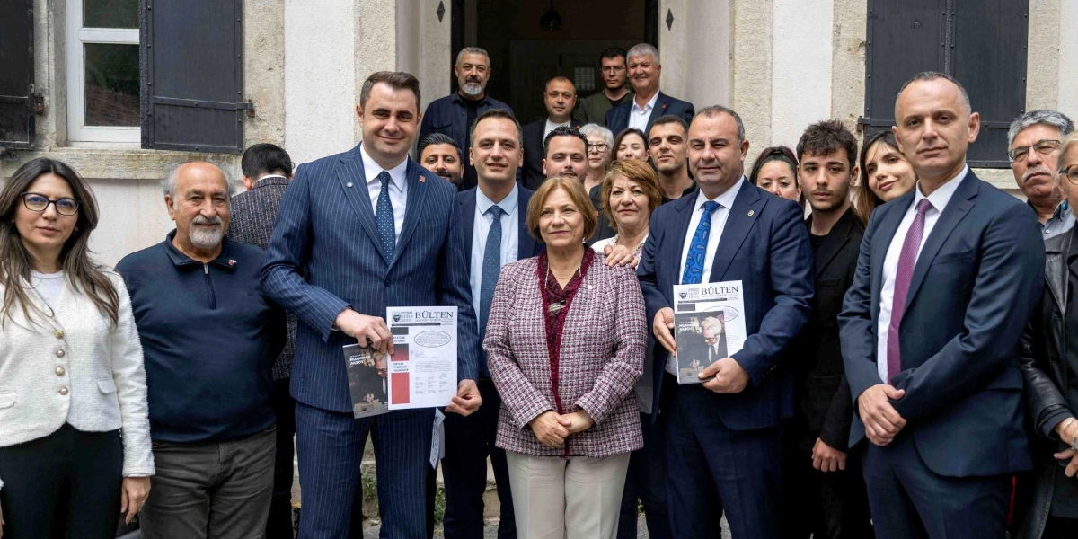 Tarihi bir binanın giriş kapısı önünde yan yana duran, aralarında takım elbiseli siyasetçilerin de bulunduğu kalabalık bir grup. Grubun ön sırasında yer alan iki erkek, ellerinde üzerinde 