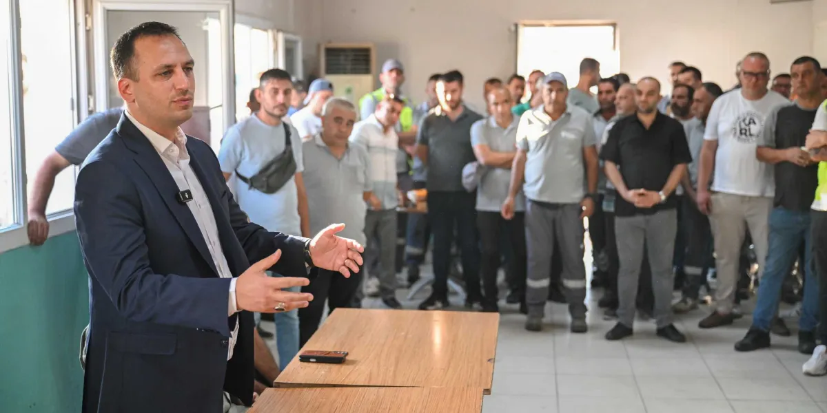Koyu renk ceketli ve beyaz gömlekli bir erkek, kapalı bir alanda önünde duran ahşap bir masanın arkasından, karşısında duran kalabalık bir gruba hitap ediyor. Gruptaki kişiler genellikle günlük kıyafetler ve iş kıyafetleri içinde, konuşmacıyı dikkatle dinliyor.