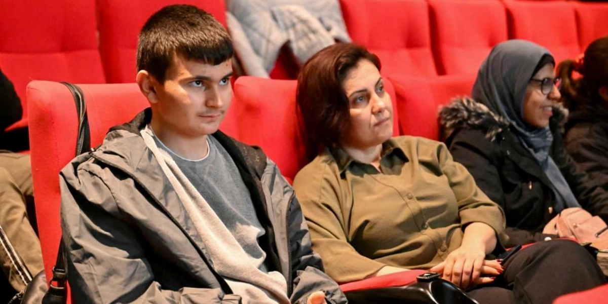 Kırmızı koltuklu bir sinema veya tiyatro salonunda yan yana oturmuş, dikkatle karşıya bakan genç bir erkek, yanında oturan orta yaşlı bir kadın ve onun yanında başörtülü bir kadın izleyici.