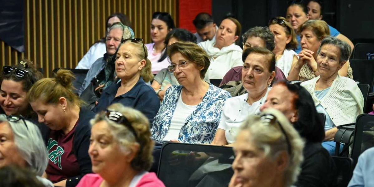 Konferans salonunda oturan, çoğunluğu kadınlardan oluşan bir izleyici grubu, dikkatle bir konuşmacıyı dinliyor. Katılımcılar farklı yaş gruplarından olup, gündelik kıyafetler içinde sıralı koltuklarda oturuyorlar. Arka planda dikey ahşap panellerden oluşan bir duvar dekoru bulunuyor.